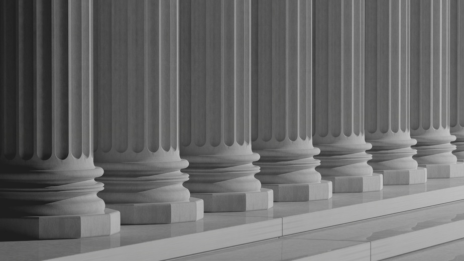A row of classic stone columns with intricate detailing, standing on a polished floor. The repetitive pattern creates a sense of symmetry and architectural grandeur. The grayscale tones add a timeless and elegant feel.