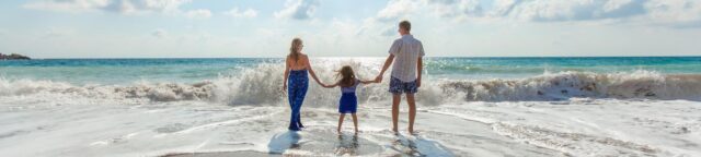 A family of three, with a child in the center, stands at the edge of the ocean, holding hands. The sky is partly cloudy, and waves are gently rolling in. They appear to be enjoying a sunny day at the beach.