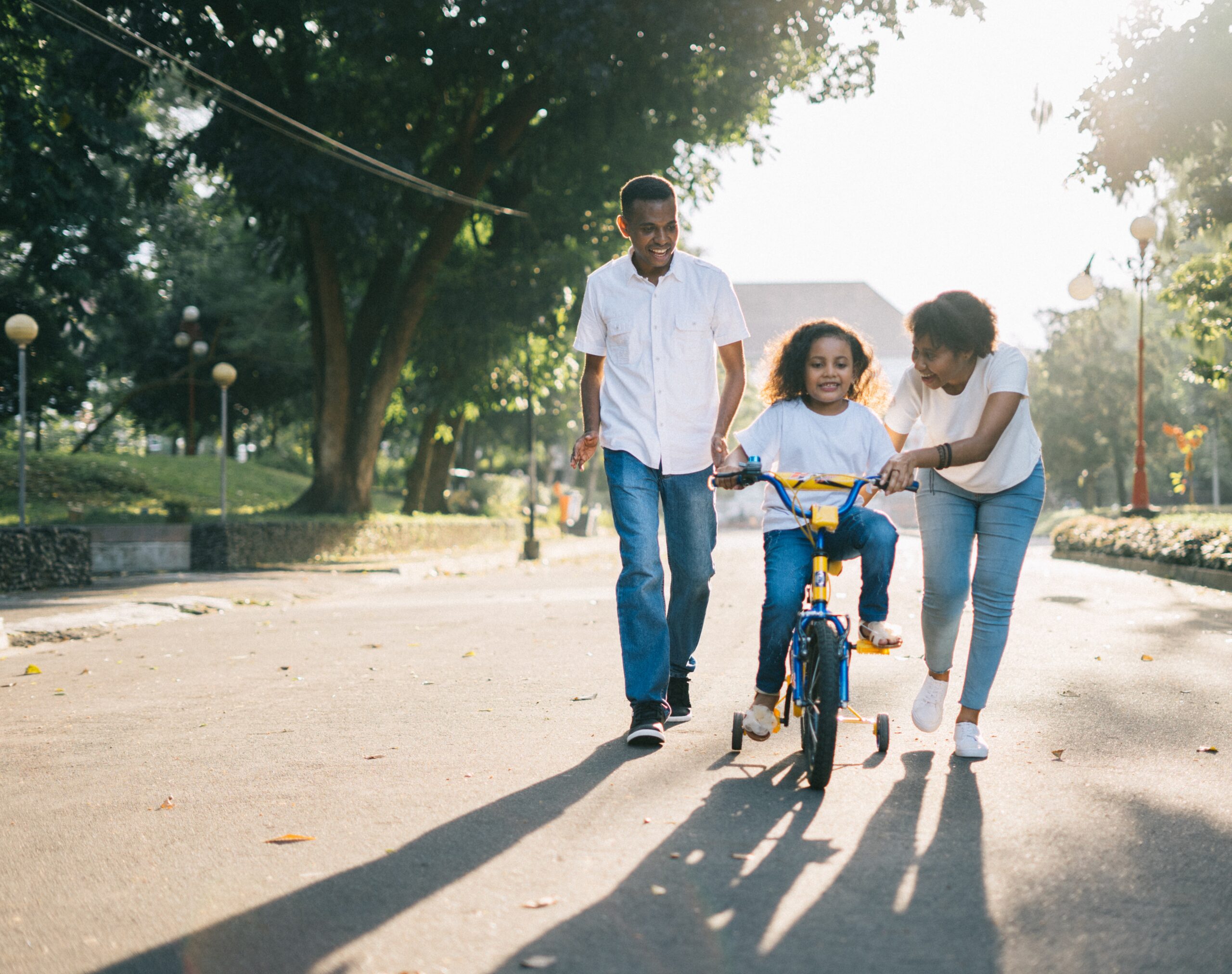 A child is learning to ride a bike with the help of two adults on a sunny day. They are on a paved path surrounded by trees, smiling and enjoying the moment.