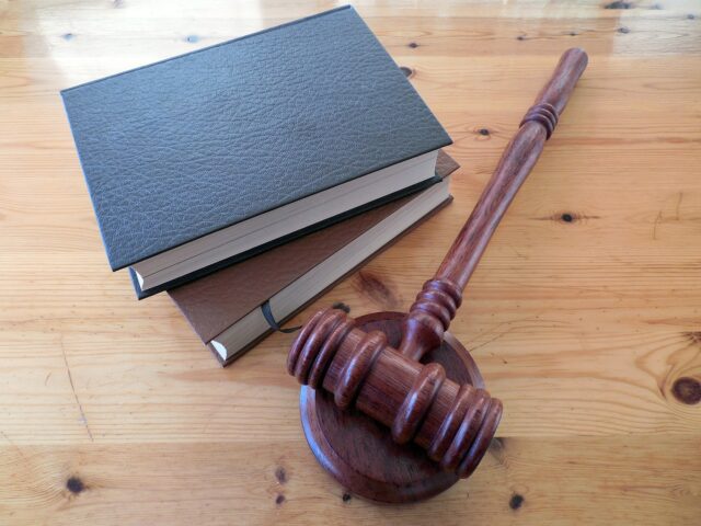 A wooden judge's gavel rests on a flat surface next to two stacked legal books with dark covers. The scene is set on a light wooden table, conveying a sense of law and justice.