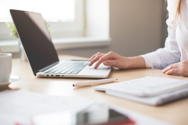A person in a white shirt uses a laptop on a wooden desk. The desk has a notebook, a pen, and a blurred cup. Sunlight filters through a window in the background, creating a bright and calm workspace.