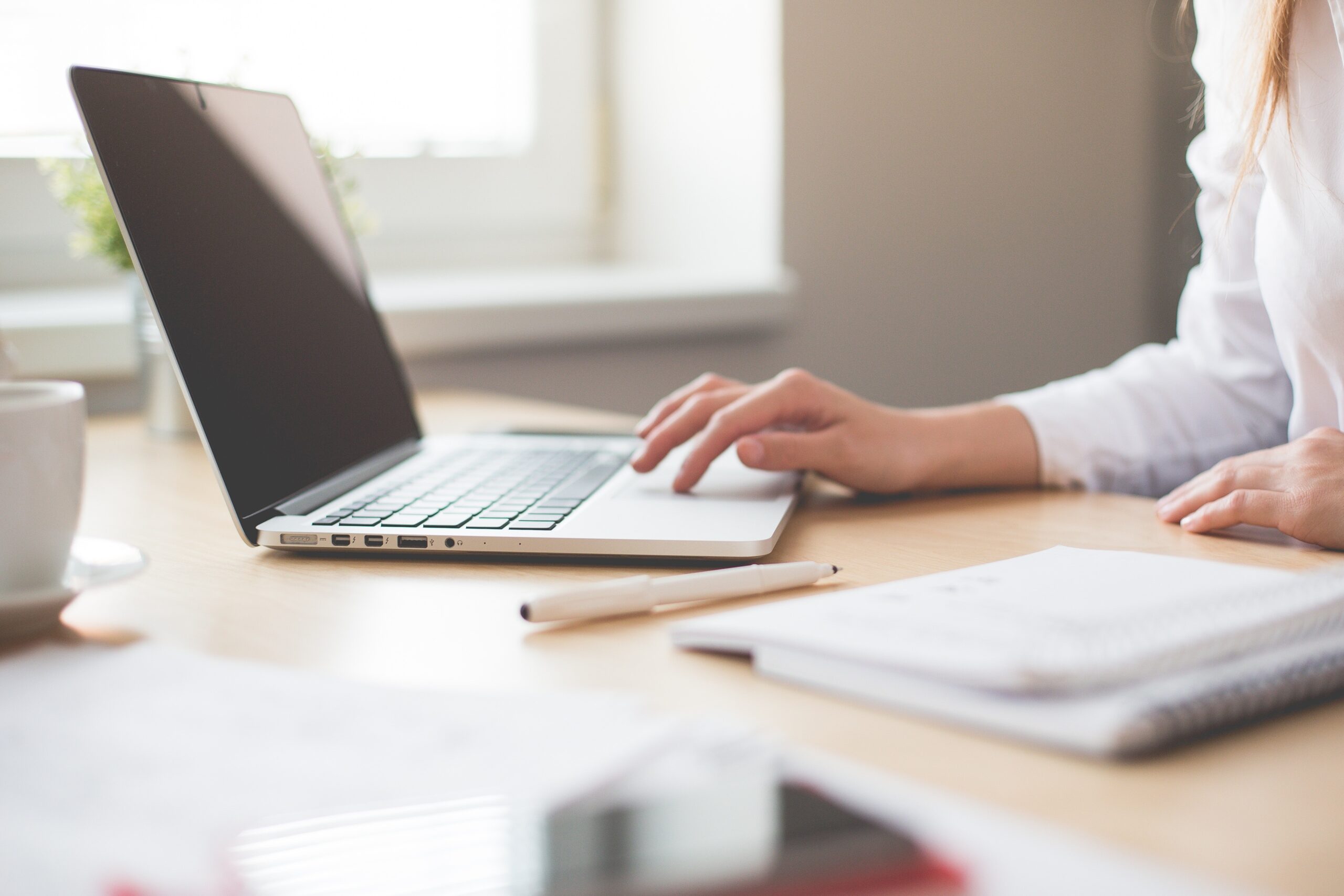 A person in a white shirt uses a laptop on a wooden desk. The desk has a notebook, a pen, and a blurred cup. Sunlight filters through a window in the background, creating a bright and calm workspace.