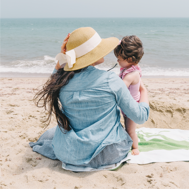 A woman in a straw hat sits on a beach with a toddler wearing a pink outfit. They face the ocean while sitting on a towel on the sand. The sky is clear and the sea is calm. The woman has her arm around the child, creating a tender moment.