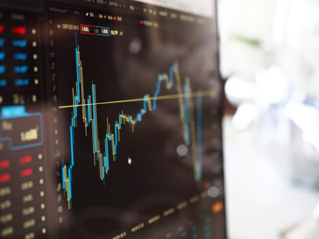 Close-up of a computer screen displaying a financial stock chart with blue and yellow lines representing market data. The background is blurred, emphasizing the focus on the fluctuating graph and numerical details on the dark screen.