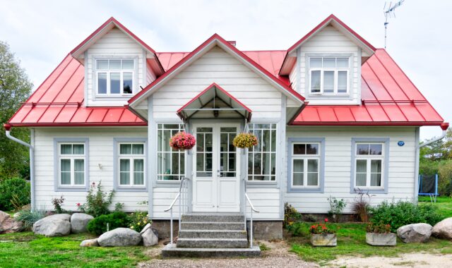 A charming home with a white facade and red roof boasts two dormer windows. The entrance features a cozy porch adorned with hanging flower baskets, while the foreground presents a delightful garden with rocks and shrubs.