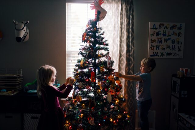 Two children, enjoying their time in a dimly lit room, decorate a Christmas tree adorned with colorful lights and ornaments. A window softly illuminates the scene, as a zebra mask and an alphabet poster hang on the wall—creating memories far removed from concerns like child custody.