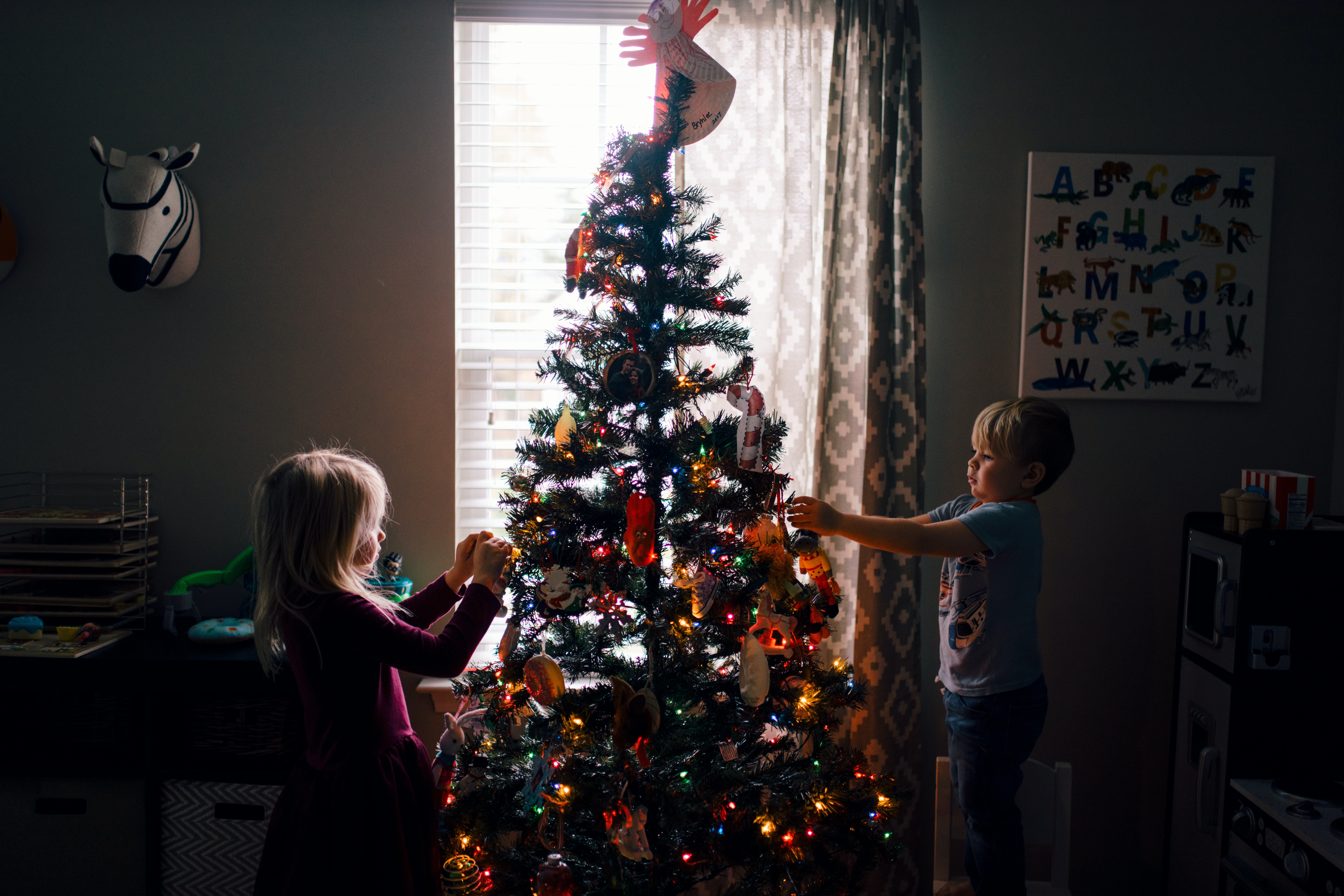 Two children, enjoying their time in a dimly lit room, decorate a Christmas tree adorned with colorful lights and ornaments. A window softly illuminates the scene, as a zebra mask and an alphabet poster hang on the wall—creating memories far removed from concerns like child custody.