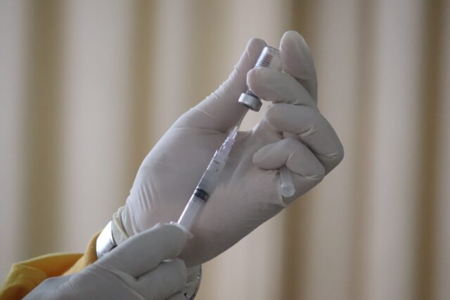 A gloved hand holds a syringe, drawing liquid from a small vial, preparing to vaccinate. The background is blurred, focusing attention on the medical procedure.
