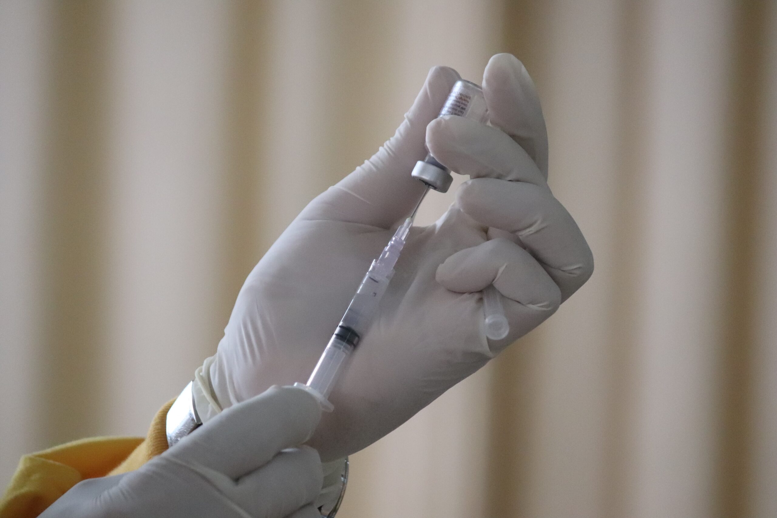A gloved hand holds a syringe, drawing liquid from a small vial, preparing to vaccinate. The background is blurred, focusing attention on the medical procedure.