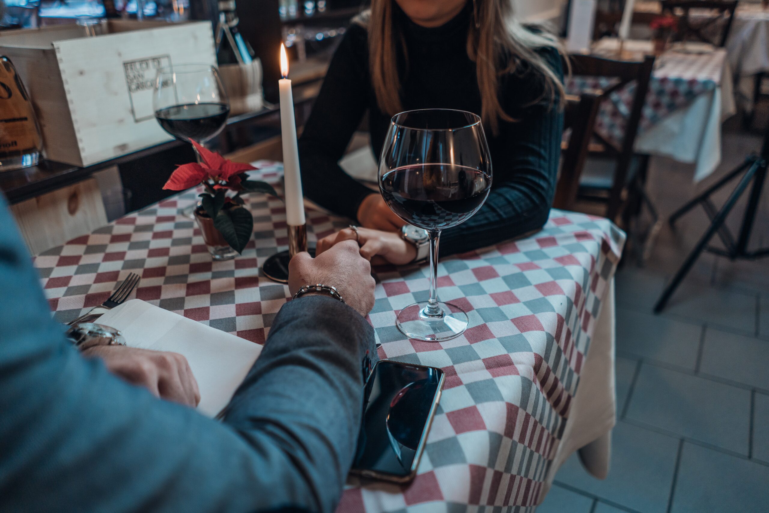 A couple sits at a table in a cozy restaurant, holding hands while navigating their date during divorce. A lit candle and two glasses of red wine rest on the checkered tablecloth, as a small red flower arrangement adds a touch of color to this emotionally complex scene.