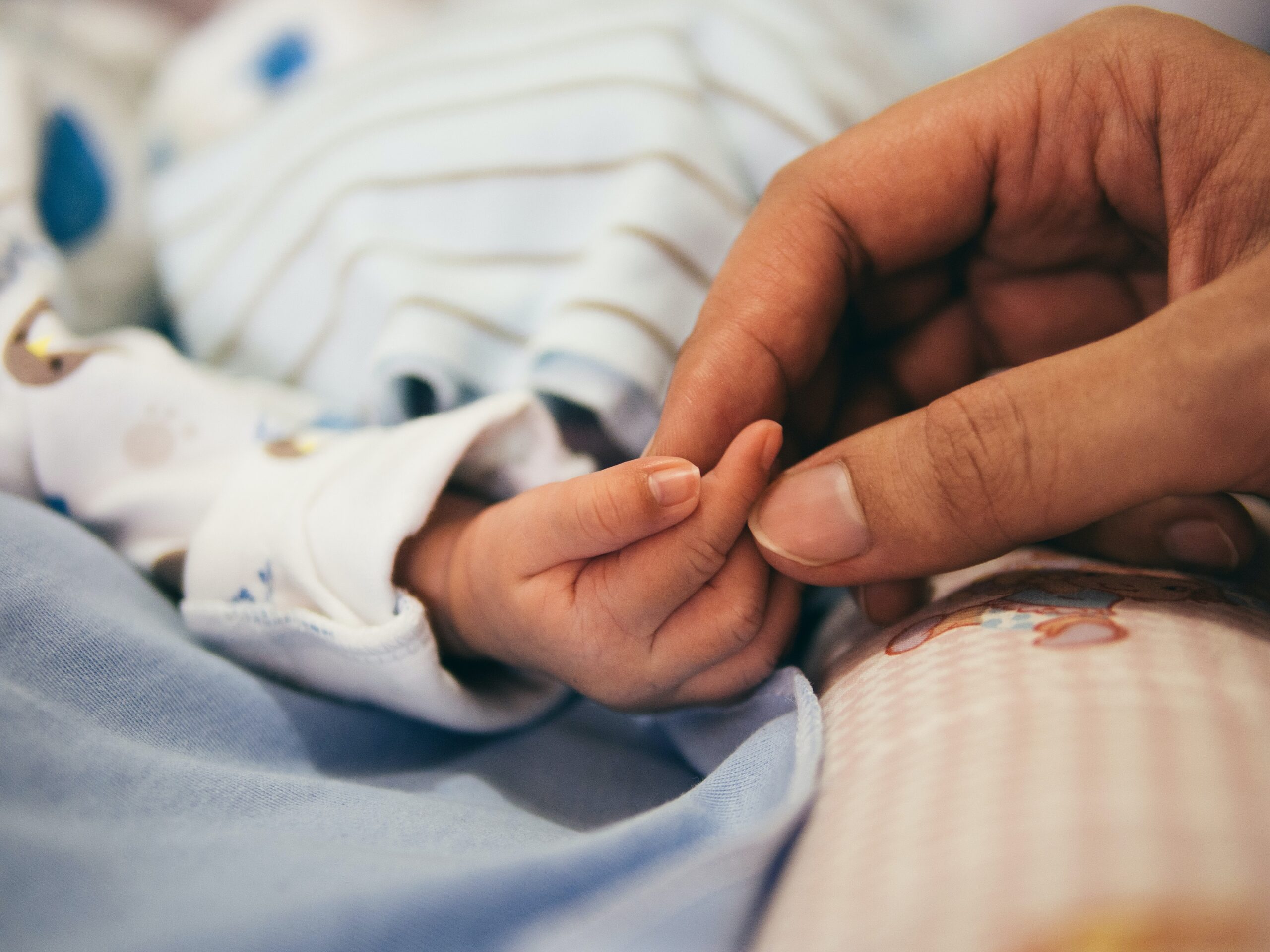 A close-up of an adult hand gently holding a baby's hand, symbolizing the journey of adoption. The baby is wearing a white onesie with a blue and brown pattern. The adult's fingers softly encircle the infant's, showcasing a tender and protective bond.