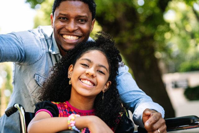 A little girl in a wheelchair enjoying and having fun while taking a selfie with her father outdoors.