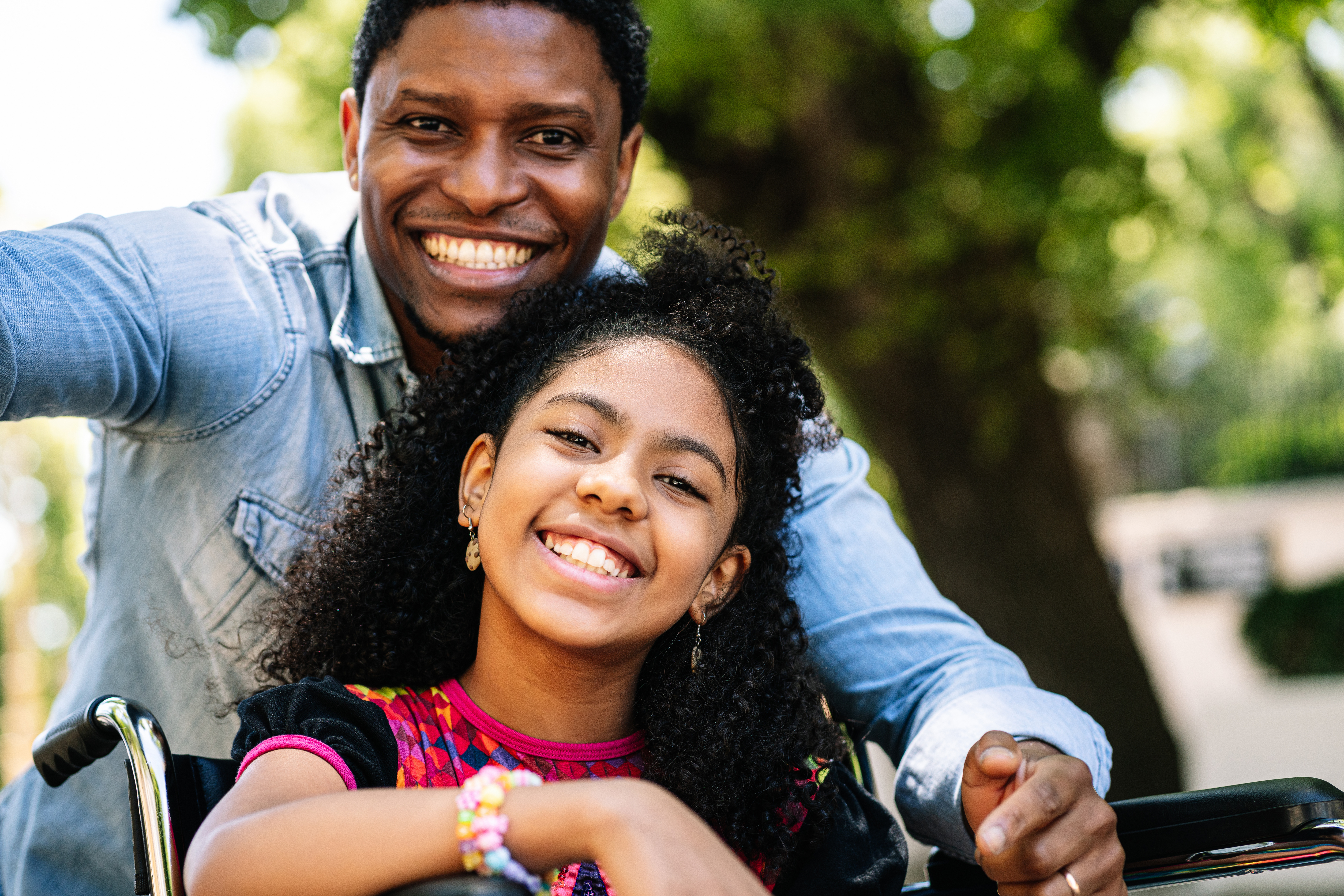 A little girl in a wheelchair enjoying and having fun while taking a selfie with her father outdoors.