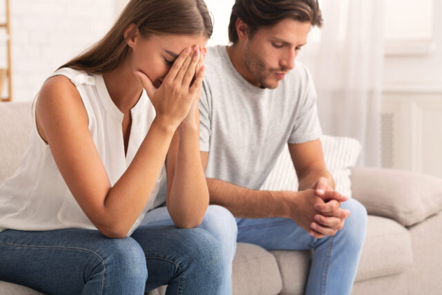 A woman and man sit on a couch, appearing upset. The woman has her hands clasped in front of her face, elbows on her knees, while the man looks downward, clasping his hands. Both wear casual clothing in a softly lit room.