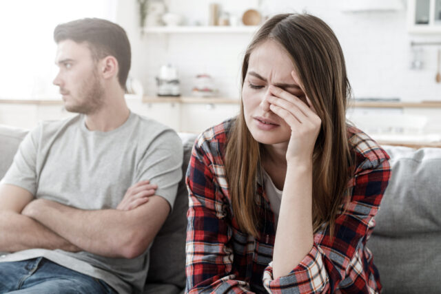 A woman in a plaid shirt sits on a couch, holding her forehead, appearing upset. A man in a gray T-shirt sits beside her, looking away with arms crossed. The background shows a kitchen area