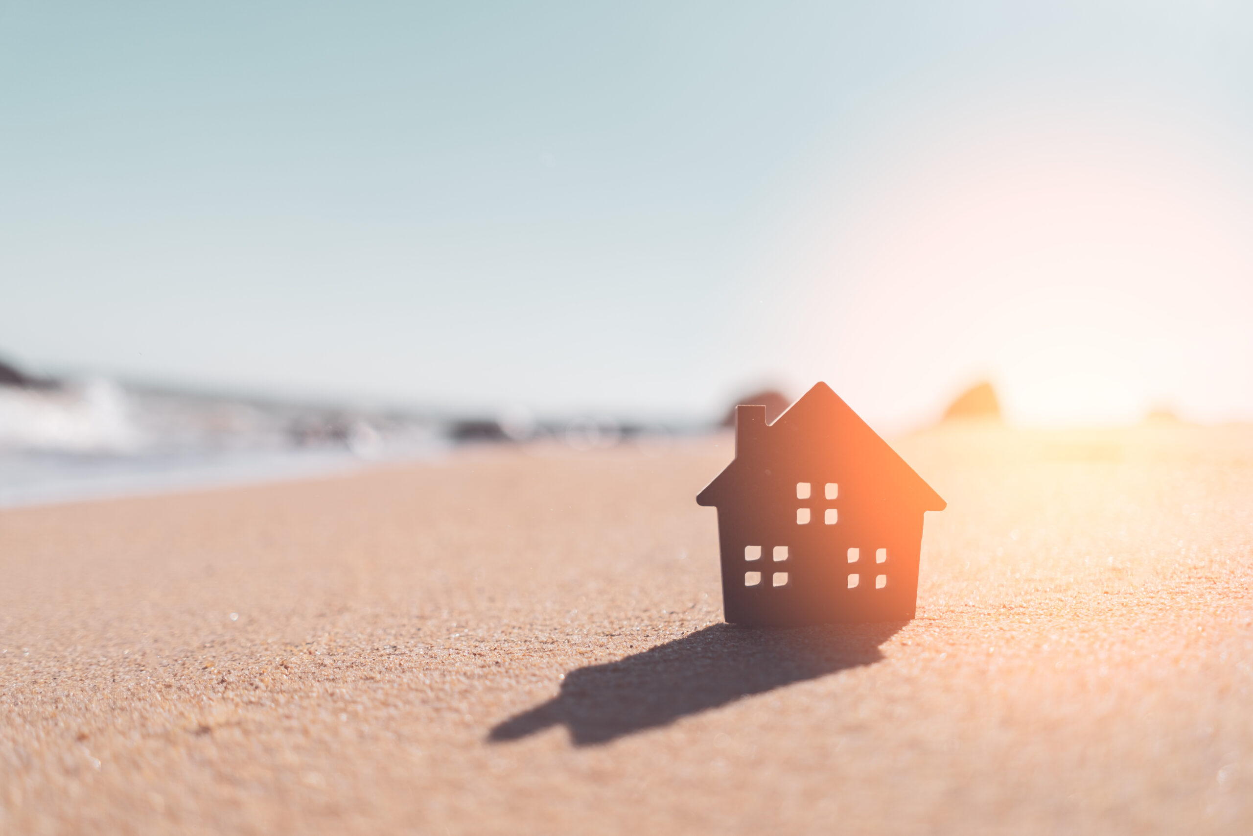 A small, dark silhouette of a vacation home stands on a sandy beach, casting a shadow. The background shows a blurred ocean and a bright, sunny sky, creating a warm and tranquil atmosphere.