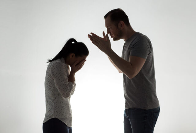 A man and a woman are silhouetted against a light background. The man, possibly an abusive spouse, stands on the right, appearing to speak loudly with his hands raised. The woman on the left faces him, her hands covering her face in distress.