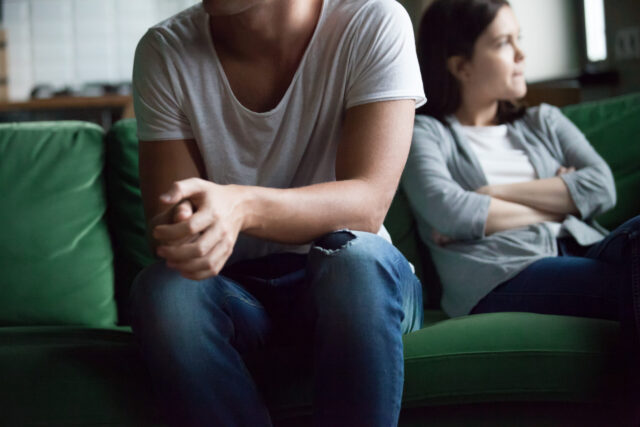 A man and woman sit on a green couch, both appearing distant. The man is in the foreground, wearing a white t-shirt and ripped jeans, with his hands clasped. The woman in the background has crossed arms and looks away. The mood seems tense.
