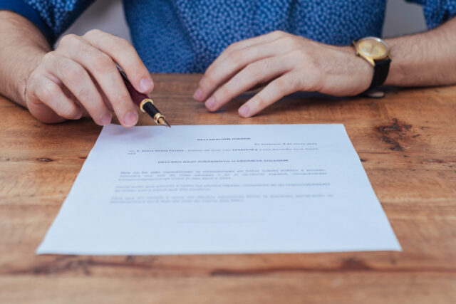 A person in a blue shirt holds a pen over a document on a wooden table, poised to sign. A gold wristwatch is visible on their left hand. The document appears to contain formal text but is mostly unreadable.