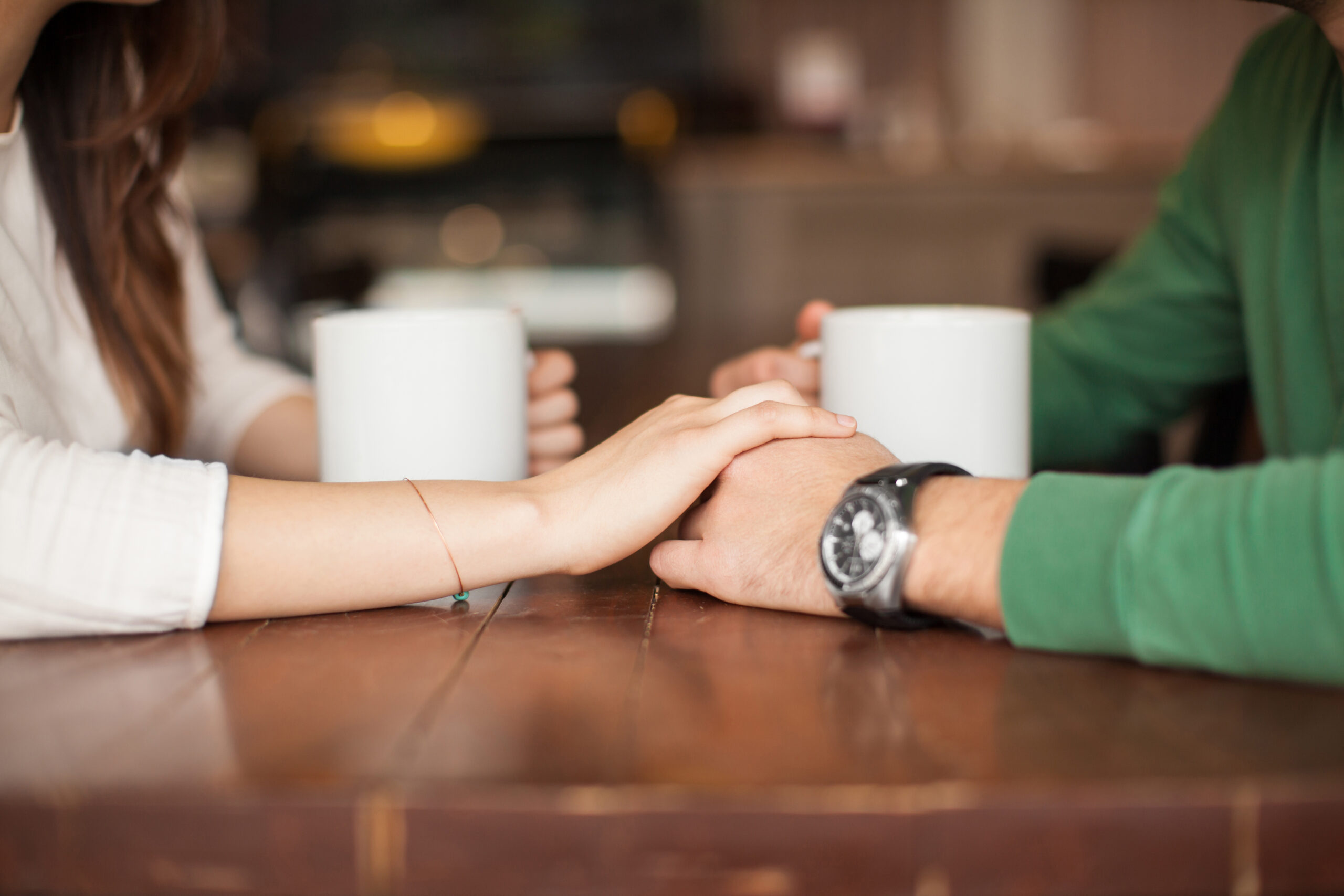 Two people sit at a wooden table, each cradling a white mug. The person on the left, in a white long-sleeve shirt, gently holds the other’s hand. The individual on the right wears a green sleeve and wristwatch—perhaps finding solace in companionship post-divorce.