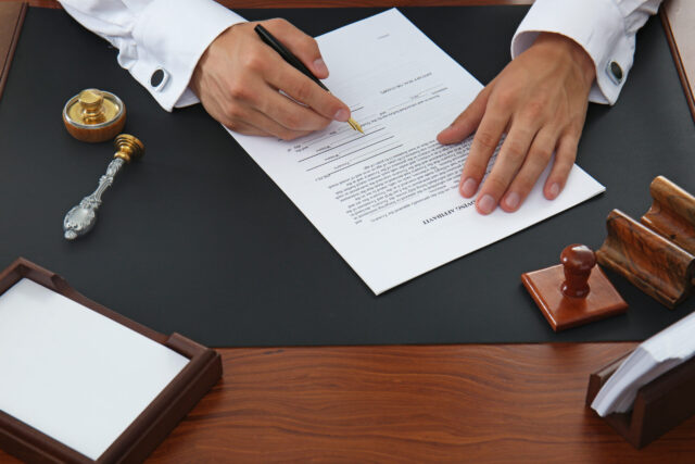 Close-up of a person signing a financial affidavit on a desk with a black mat. Nearby are a pen in a holder, a decorative key, a stamp, wooden organizers, and an empty notepad. The person wears a white shirt and is holding the black pen with precision.