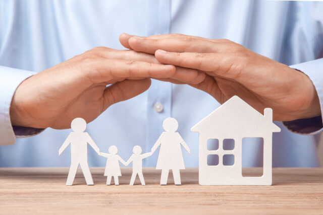 Hands forming a protective gesture over miniature cutouts of a family and a house. The family figures consist of two adults and two children, symbolizing protection and care. The setting conveys a theme of home security and insurance.