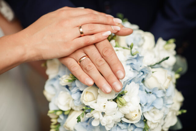 The bride and groom in wedding suits are holding together a wedding bouquet of white and lilac roses. Men's blue classic suit for men and girls white dress. Newlyweds without faces. Close up only hands